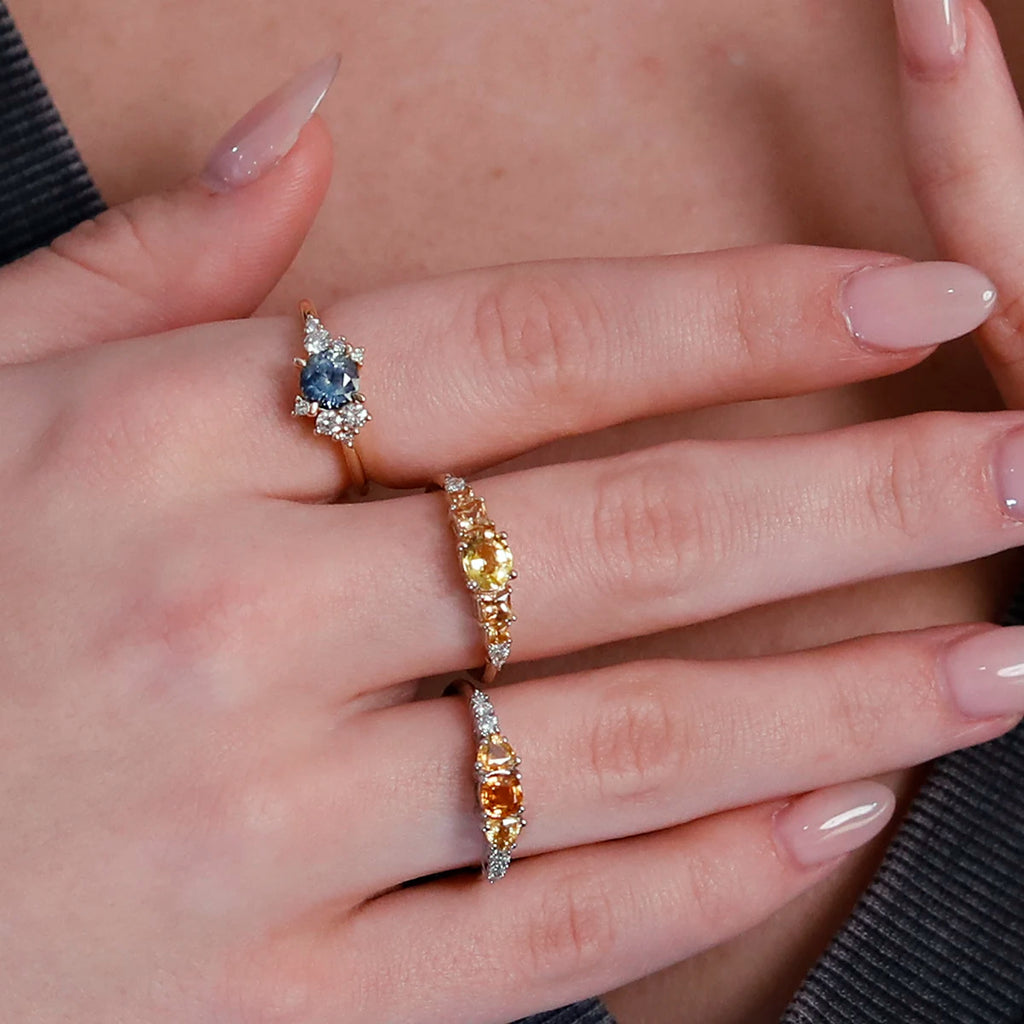 Close-up product photography of a unique engagement ring featuring a stunning round soft blue sapphire, worn on a finger with two additional gemstone rings featuring yellow and orange sapphires. The blue sapphire ring is an asymmetrical design by Justine Quintal, while the yellow and orange sapphire rings are exquisite creations from Ruby Mardi. This combination showcases a vibrant and elegant display of fine jewelry.