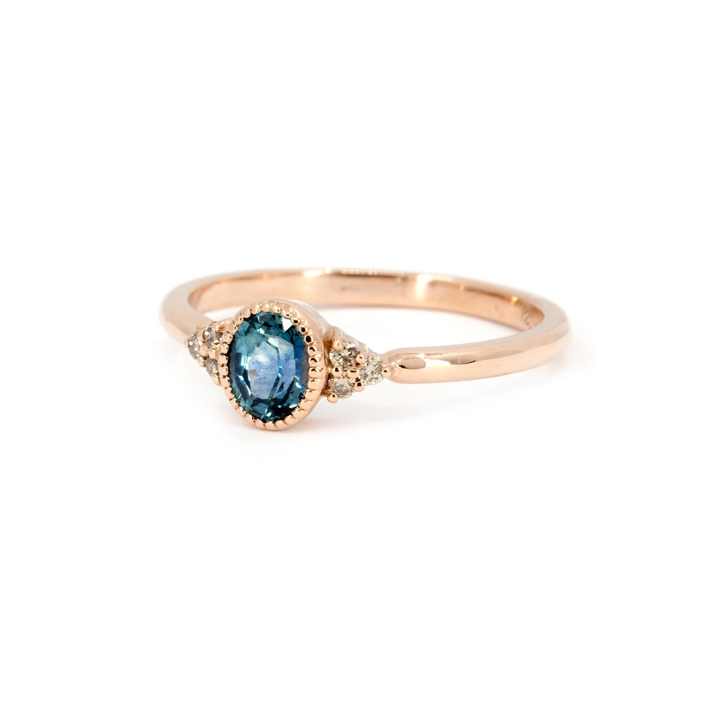 Side view of a rose gold engagement ring featuring an oval teal sapphire and champagne diamond accents, photographed on a white background.