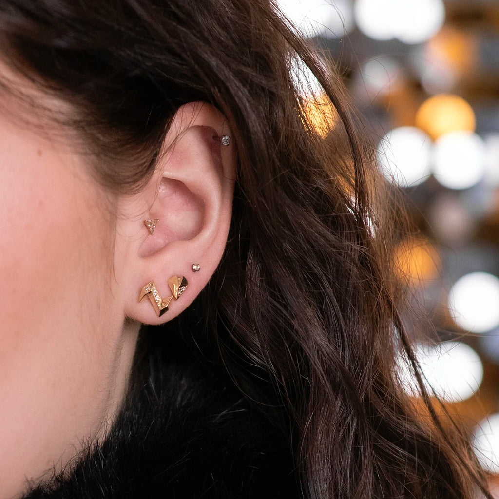 Close-up of a woman's ear adorned with modern angular heart-shaped yellow gold earrings featuring four small diamonds, paired with a small gold ball earring and an edgy piece by Bena Jewelry. The woman has long, wavy brown hair against a festive, sequin background.