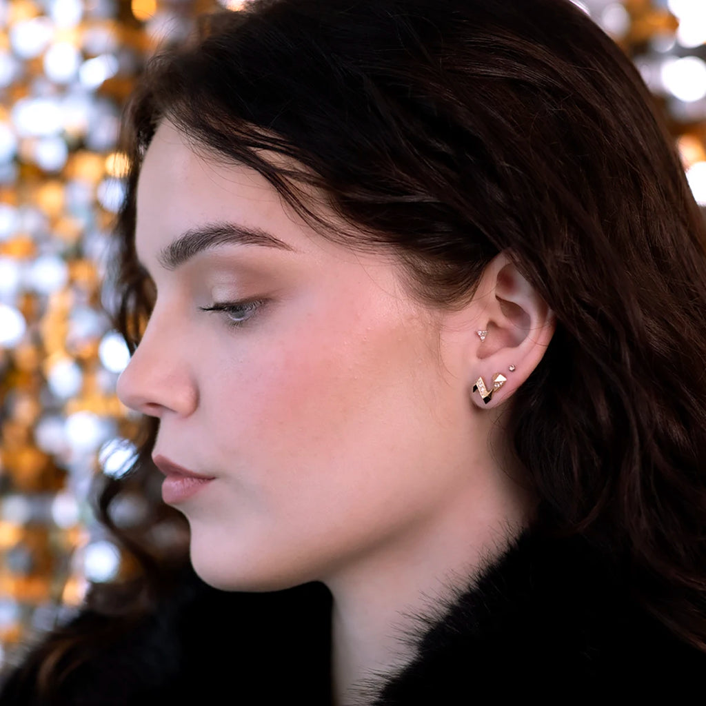 A close-up side profile of a person wearing elegant gold earrings with a geometric design, paired with minimalist stud piercings. The warm background lighting enhances the modern and sophisticated look of the jewelry.