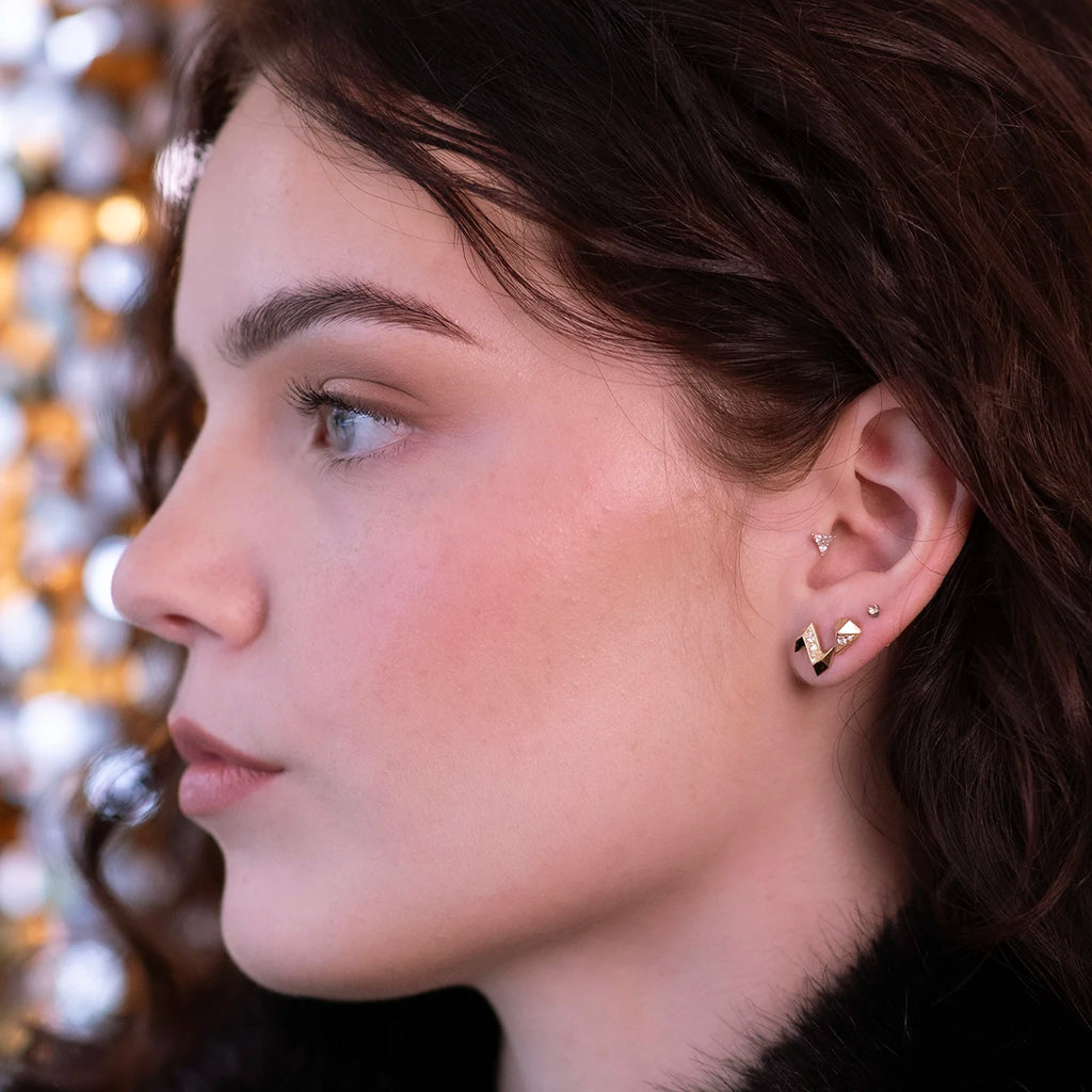 A close-up side profile of a person wearing elegant gold earrings with a geometric design, paired with minimalist stud piercings. The warm background lighting enhances the modern and sophisticated look of the jewelry.