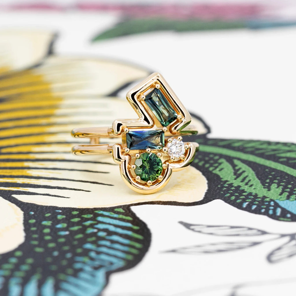 A Canadian designer ring like you've never seen before, photographed against a colorful background that recalls the color of the gemstones in the ring: blue, green, white and yellow. The ring consists of a stunning geometric composition. A rounded yellow gold band encircles 4 gemstones: a white diamond, a round green sapphire, and two bicolored sapphire baguettes. This unique creation by designer Erica Leal is available at Ruby Mardi fine jewelry.
