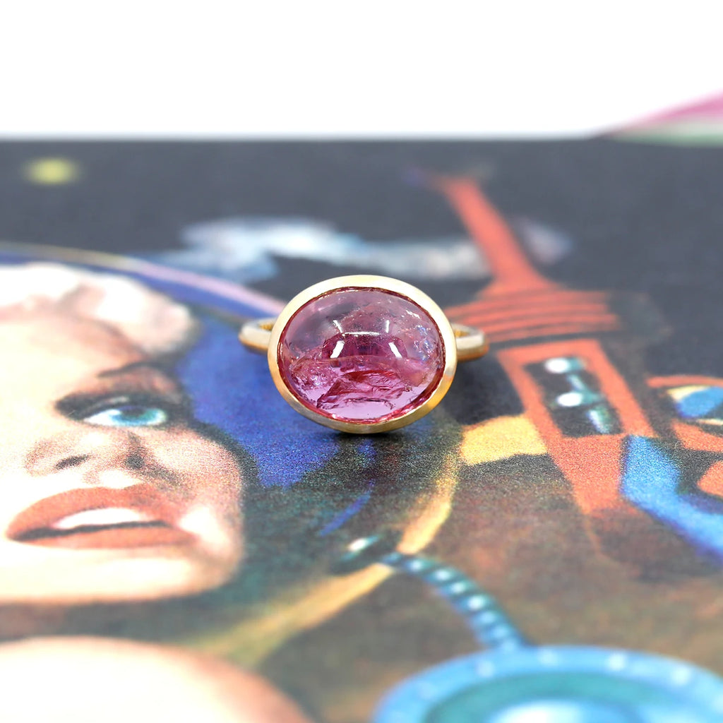 A ring that looks unatural is seen photographed on a image evoking space and starships. The big cocktail ring features a huge pink ruby cabochon bezel set in yellow gold. 
