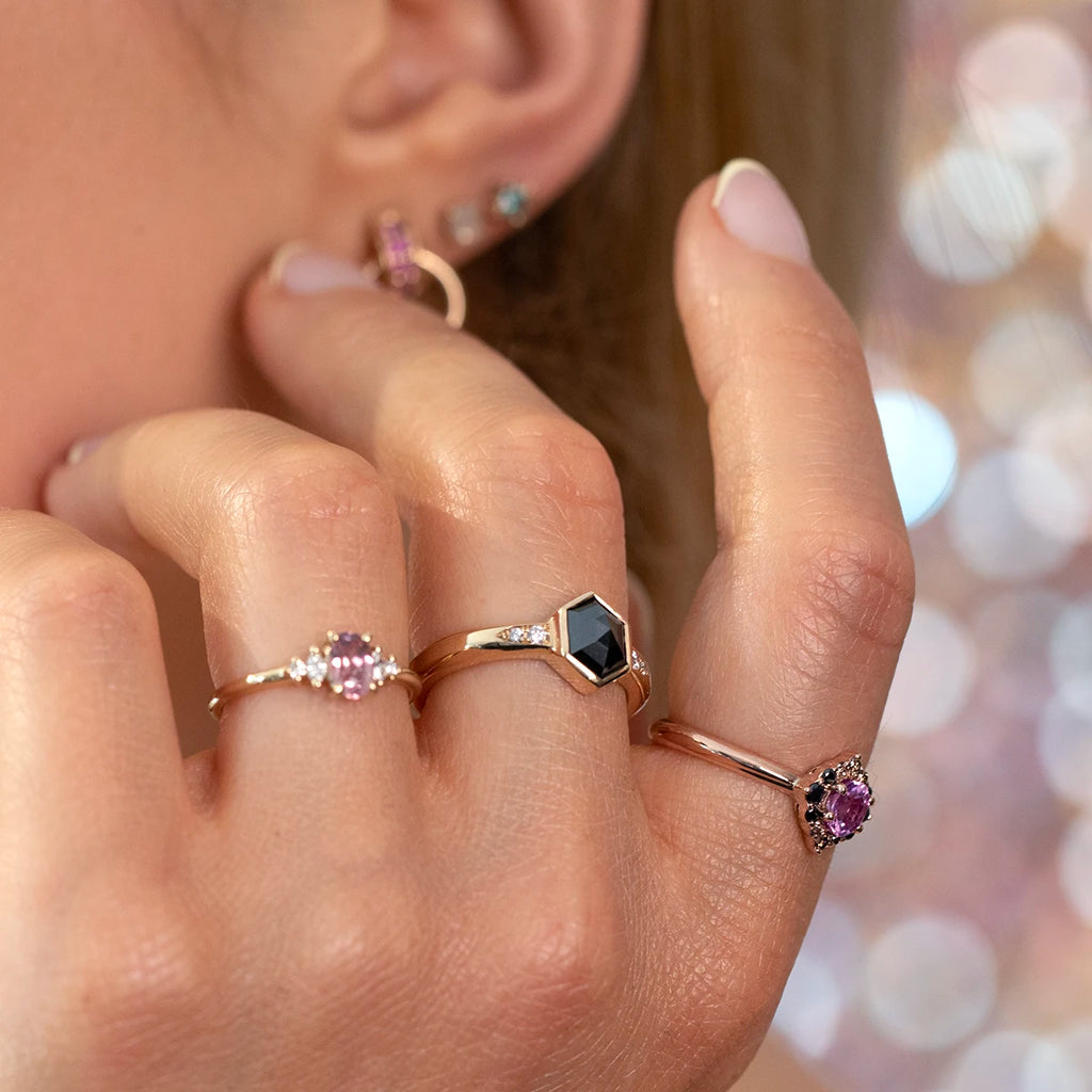 Hand wearing a trio of designer gemstone rings in rose gold: an oval pink sapphire engagement ring with diamond side stones, a bold hexagonal black diamond ring with geometric lines, and a vintage-inspired pink sapphire halo ring accented with black diamonds. Artistic fine jewelry styling, photographed worn for Ruby Mardi’s modern bridal and statement ring collection.