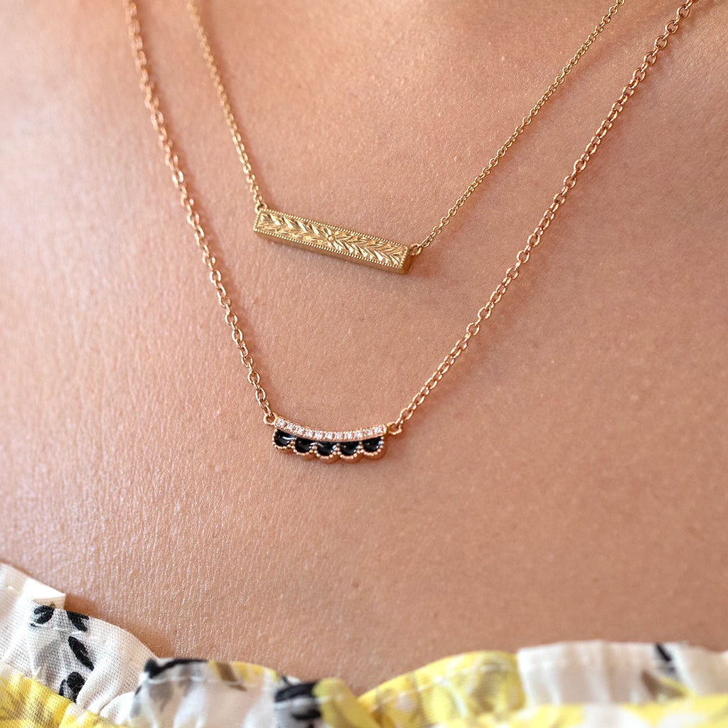 Ruby Mardi necklace in 14k rose gold by Canadian designer Emily Gill, featuring a curved pendant with black enamel scallop detail and natural diamonds. Shown worn layered with another gold pendant necklace for an elegant and modern look.