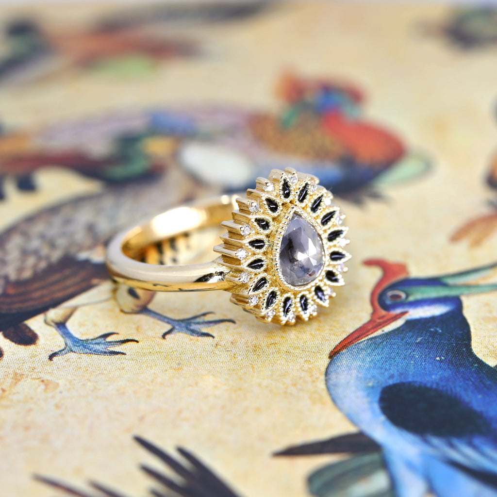  Close-up view of an original yellow gold ring photographed on an illustration showing a blue bird. The ring features a superb rose cut salt-and-pepper diamond (several facets on the stone's surface) and the diamond's inclusions are clearly visible in the photo.  Surrounding the central gem is a fine enamel work: there are 16 small black drops set off by natural round diamonds. A unique work by independent designer Emily Gill.