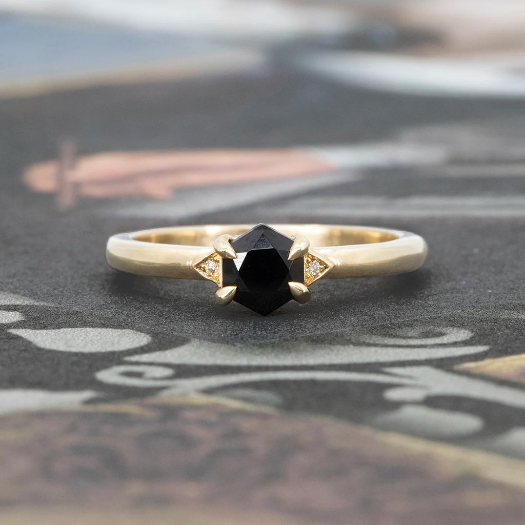 Close-up of the Luna Hex ring by Liane Vaz, featuring a hexagonal black spinel set in hand-carved yellow gold prongs, flanked by tiny triangular-set champagne diamonds, displayed on an artistic printed background.
