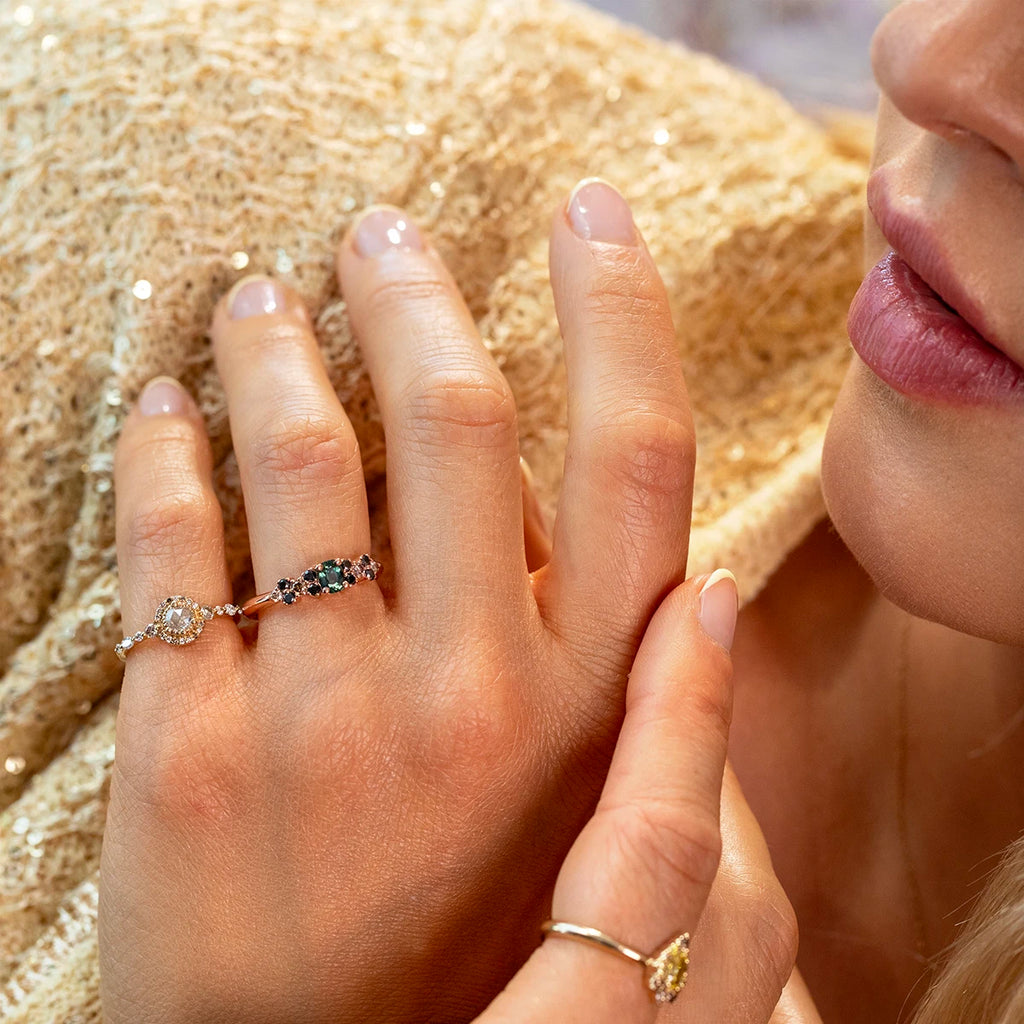 Close-up of a woman’s hand wearing three rings, including a gold ring with teal sapphires and black diamonds in a linear cluster design. Designer jewelry by Ruby Mardi, Montreal, Canada.
