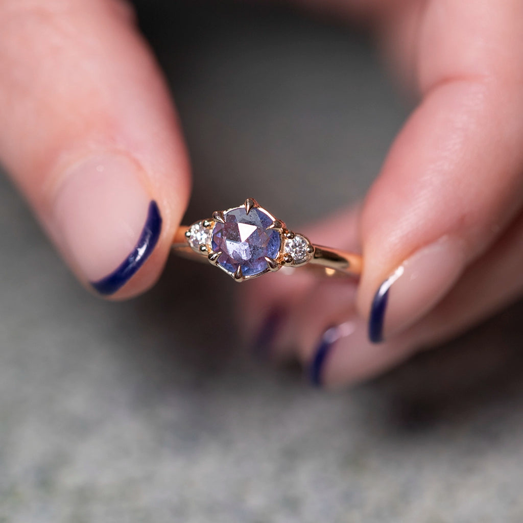 Close-up of the Blue Luna ring by Yuliya Chorna for Ruby Mardi in Montreal, Canada. Solid 14k yellow gold with a natural rose-cut round blue sapphire and Canadian diamonds in a three-stone layout. Held between fingers to highlight the handcrafted setting and the subtle glow of the sapphire.