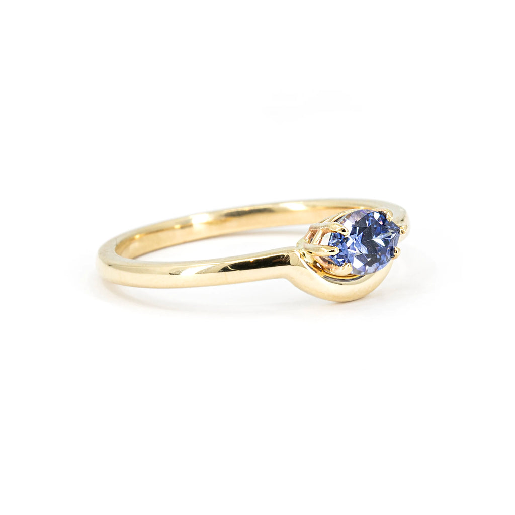 A side view on a white background of a lovely engagement ring or right hand ring featuring a marquise-cut blue sapphire with a ring encircling the gemstone to form a half-moon. This creation is a one-of-a-kind piece of jewelry from independent American brand In the Light of Day Jewelry. This unique ring is available only at Ruby Mardi, a Canadian jewelry store located in Montreal's Little Italy.