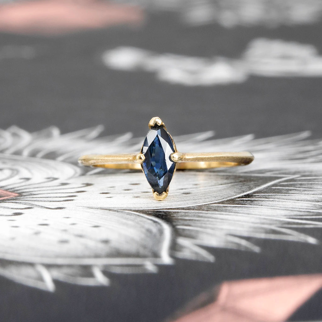 Marquise blue sapphire solitaire ring with knife-edge yellow gold band and textured finish, fine jewelry by Ruby Mardi, displayed on illustrated background.