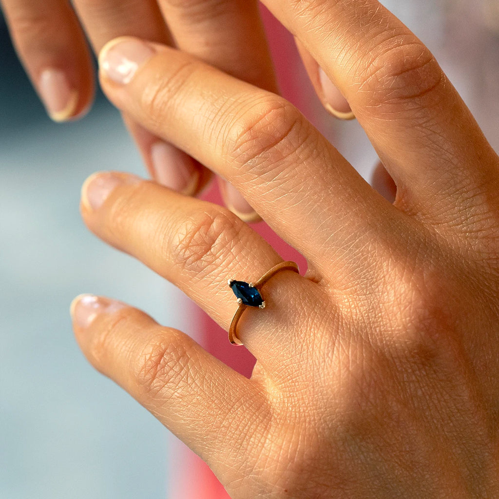 Close-up of hand wearing marquise blue sapphire solitaire ring with knife-edge yellow gold band and textured finish, fine jewelry by Ruby Mardi.