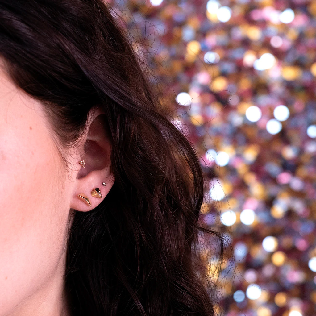 Close-up of a woman's ear adorned with modern angular heart-shaped yellow gold earrings featuring four small diamonds, paired with a small gold ball earring and an edgy piece by Bena Jewelry. The woman has long, wavy brown hair against a festive, sequin background.