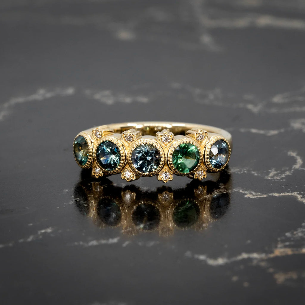 A vibrant gold ring featuring round teal sapphires in milgrain bezel settings, accented with delicate diamond details. Designed by Bramston Goldsmithing, available at Ruby Mardi in Montreal.