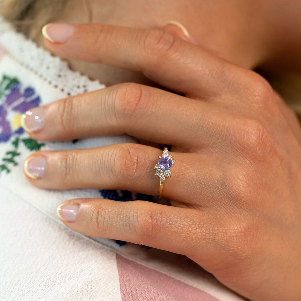 A gold ring with a round purple sapphire center stone and asymmetrical diamond accents, worn on a hand with light pink manicured nails and floral-embroidered clothing visible in the background.