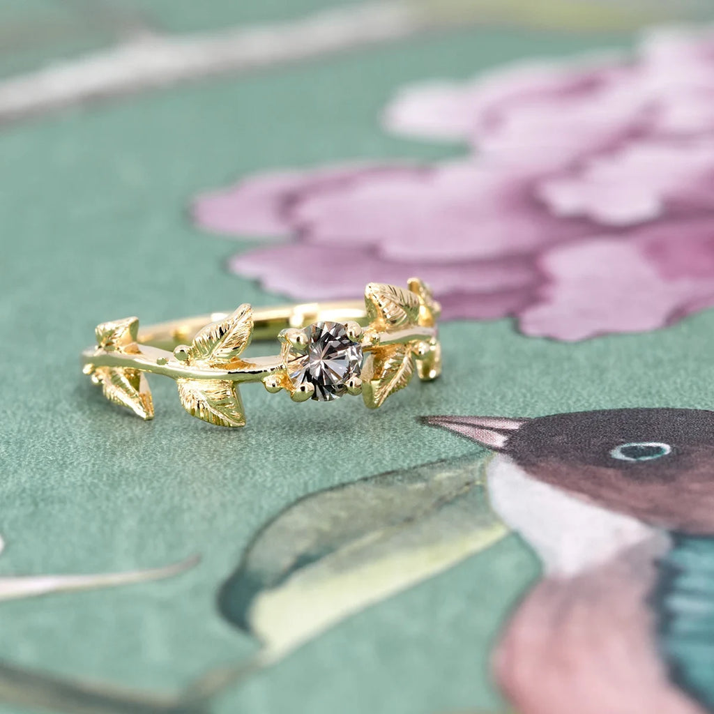 14k gold engagement ring with leaf motif and champagne sapphire, photographed on a floral green background. Nature-inspired design by Meg Lizabet, available at Ruby Mardi in Montreal.