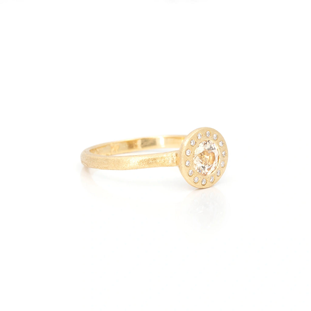 Yellow gold signet ring with a central champagne sapphire (of a really light pink color) and 14 small round brilliant cut diamonds bezel set around the central gemstone. The ring is photographed from the side on a white background, and we can see the rough texture of the gold band. It’s a one-of-a-kind jewel handmade in Toronto by Anouk Jewelry and available only at boutique Ruby Mardi in Montreal.