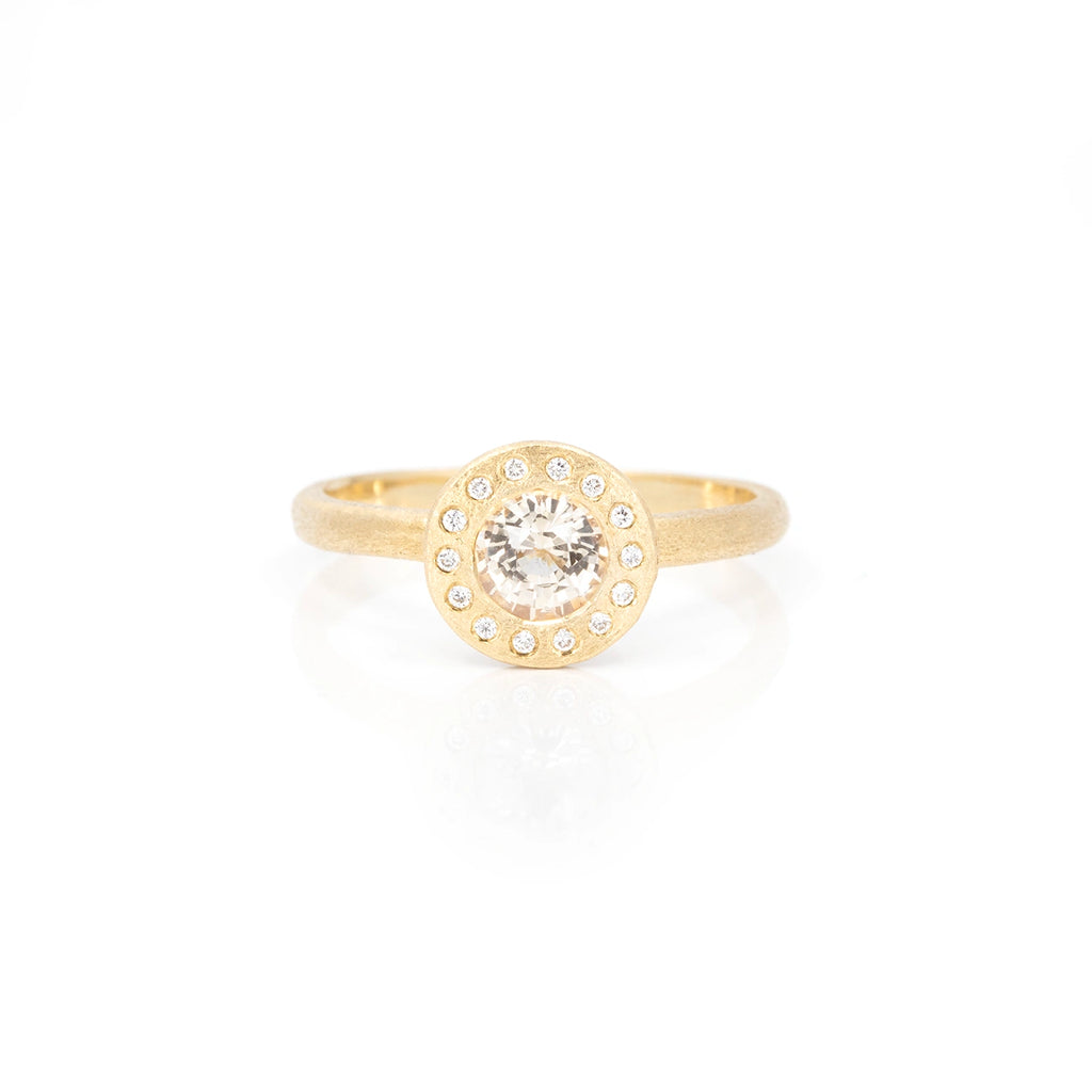 A yellow gold ring with a rough finition is photographed on a white background. The ring presents a round face with a central bezel set champagne sapphire (almost transparent gemstone) and is surrounded by 14 small natural diamonds. This jewel would be perfect as an engagement ring, or right hand ring. It is one-of-a-kind and is only available at Ruby Mardi, a fine jewelry store in Montreal.