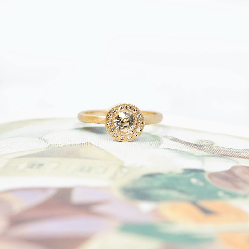 A yellow gold ring with a rough finition is photographed on an artistic background. The ring presents a round face with a central bezel set light pink gemstone (a champagne sapphire) and is surrounded by 14 small round brilliant natural diamonds. This jewel would be perfect as a bridal ring, or right hand ring. It is one-of-a-kind and is only available at Ruby Mardi, a fine jewelry store in Montreal’s Little Italy.