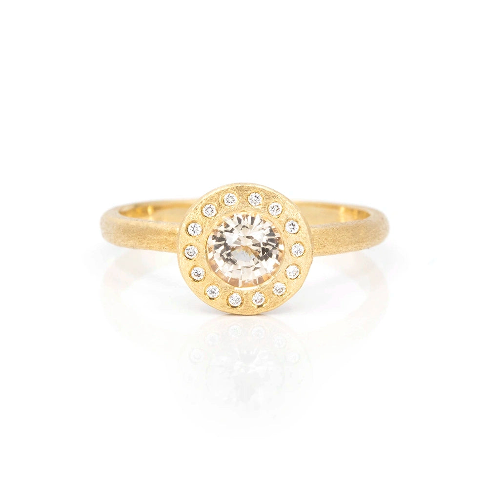 A yellow gold ring with a rough finition is photographed on a white background. The ring presents a round face with a central bezel set champagne sapphire (almost transparent gemstone) and is surrounded by 14 small natural diamonds. This jewel would be perfect as an engagement ring, or right hand ring. It is one-of-a-kind and is only available at Ruby Mardi, a fine jewelry store in Montreal.