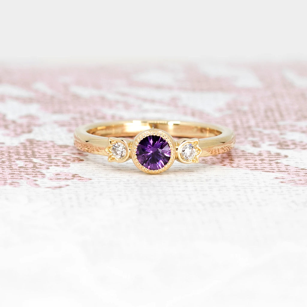 The front view of this engagement ring with a purple colored gem, a natural sapphire in a rectangular shape and surrounded by two small round diamonds. With a handmade pattern this one-of-a-kind wedding abgue is made by independent designer Bramston Goldsmithing of Canada in collaboration with Ruby Mardi jewelry.