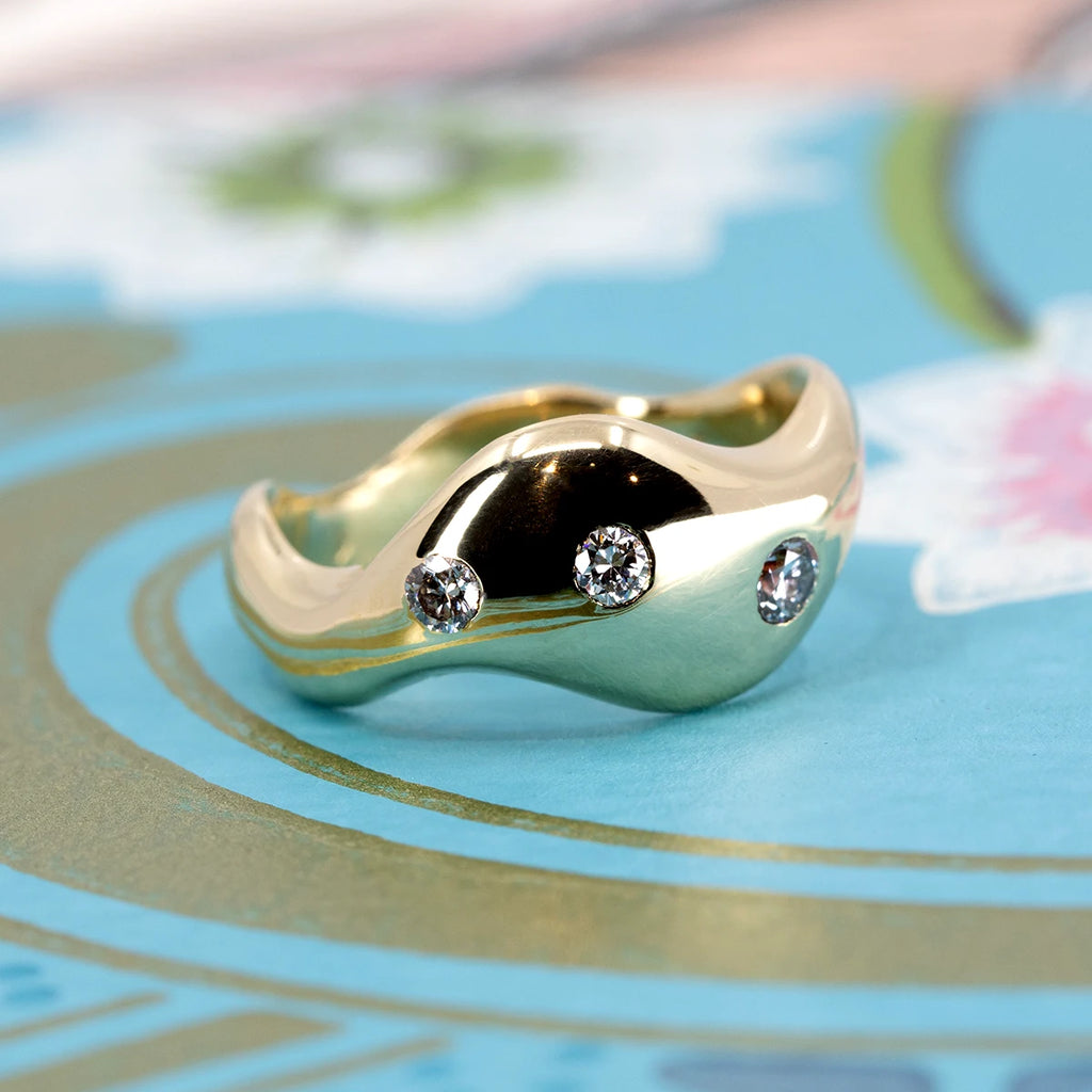 High-polish chunky gold ring with fluid, organic curves and three lab-grown diamonds, photographed on a colorful background. Available at Ruby Mardi, Montreal.