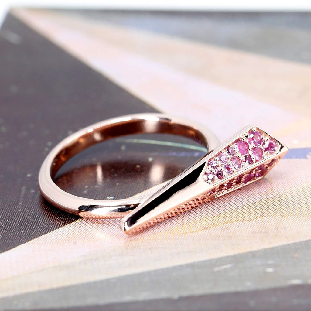  A bird's-eye view of a rose gold ring set with pink sapphires. The ring is delicate, resembling an arrow in volume, with a small point at one end, and the other end larger. This unique ring is photographed against an abstract background of different colors. It is a creation of independent Canadian brand Bena Jewelry. This modern, unisex piece of jewelry is available at Ruby Mardi, Canada's best jewelry store with its vast selection of unique designer jewelry.