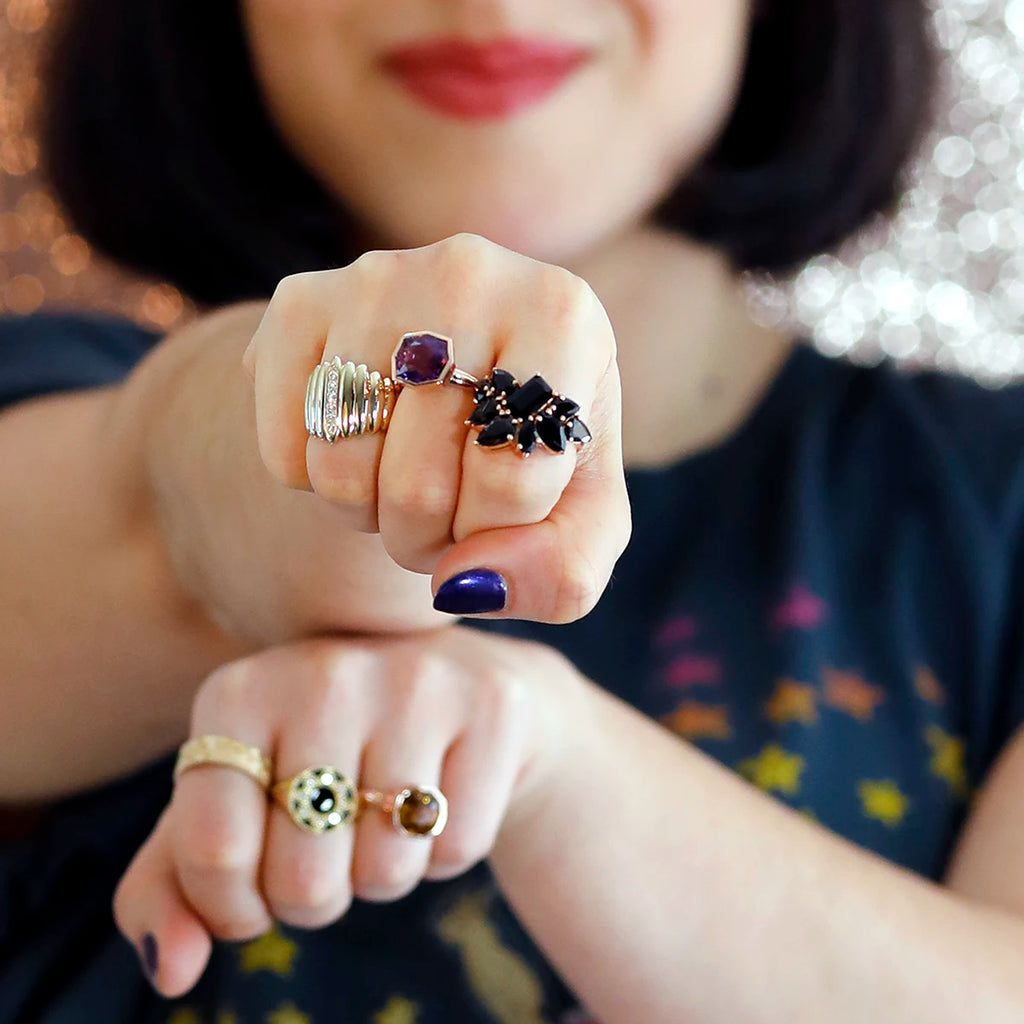 Close-up of a woman's hands adorned with statement rings, including a bold black tourmaline cluster ring in rose gold and an amethyst bezel-set ring by Bena Jewelry. Her nails are painted dark purple, and she wears additional gold rings with intricate designs. The blurred background features sparkling lights.