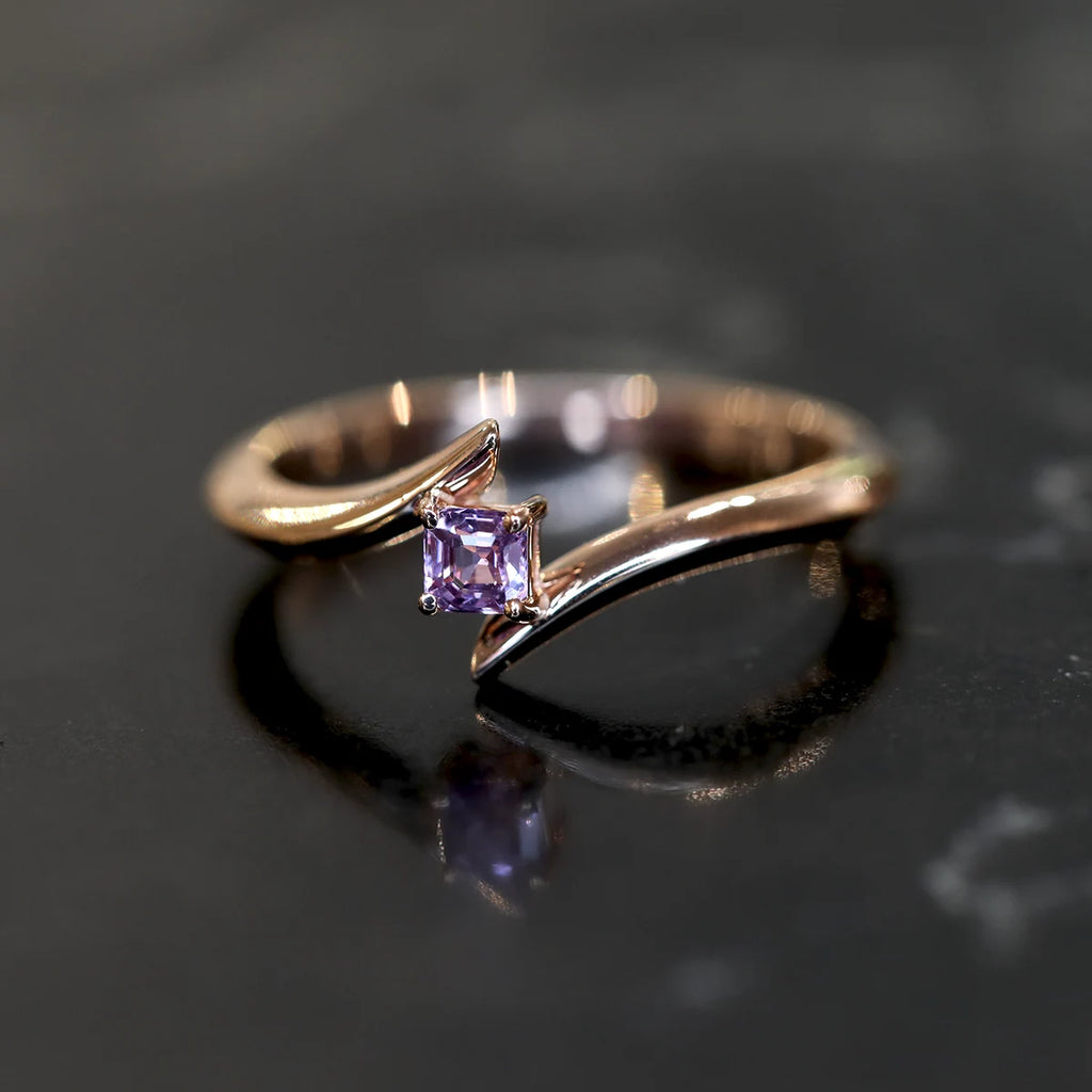 Twisted rose gold engagement ring with an emerald cut purple-pink sapphire and a twisted band, photographed on a black background, handmade in Montreal by local jewelry brand Ruby Mardi.
