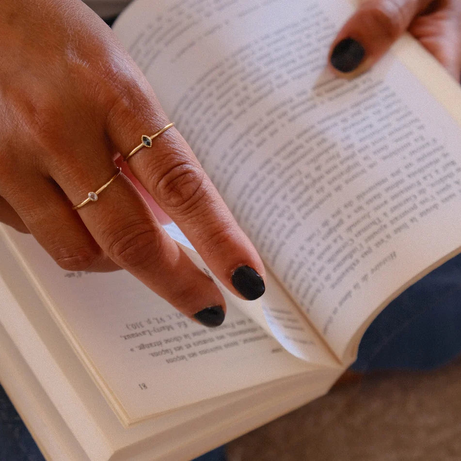 Women's hand wearing gold rings with diamond and sapphire while leafing through a book. Made by independent Canadian jewelry designer Émigé and available for sale at the Ruby Mardi jewelry store in Montreal.