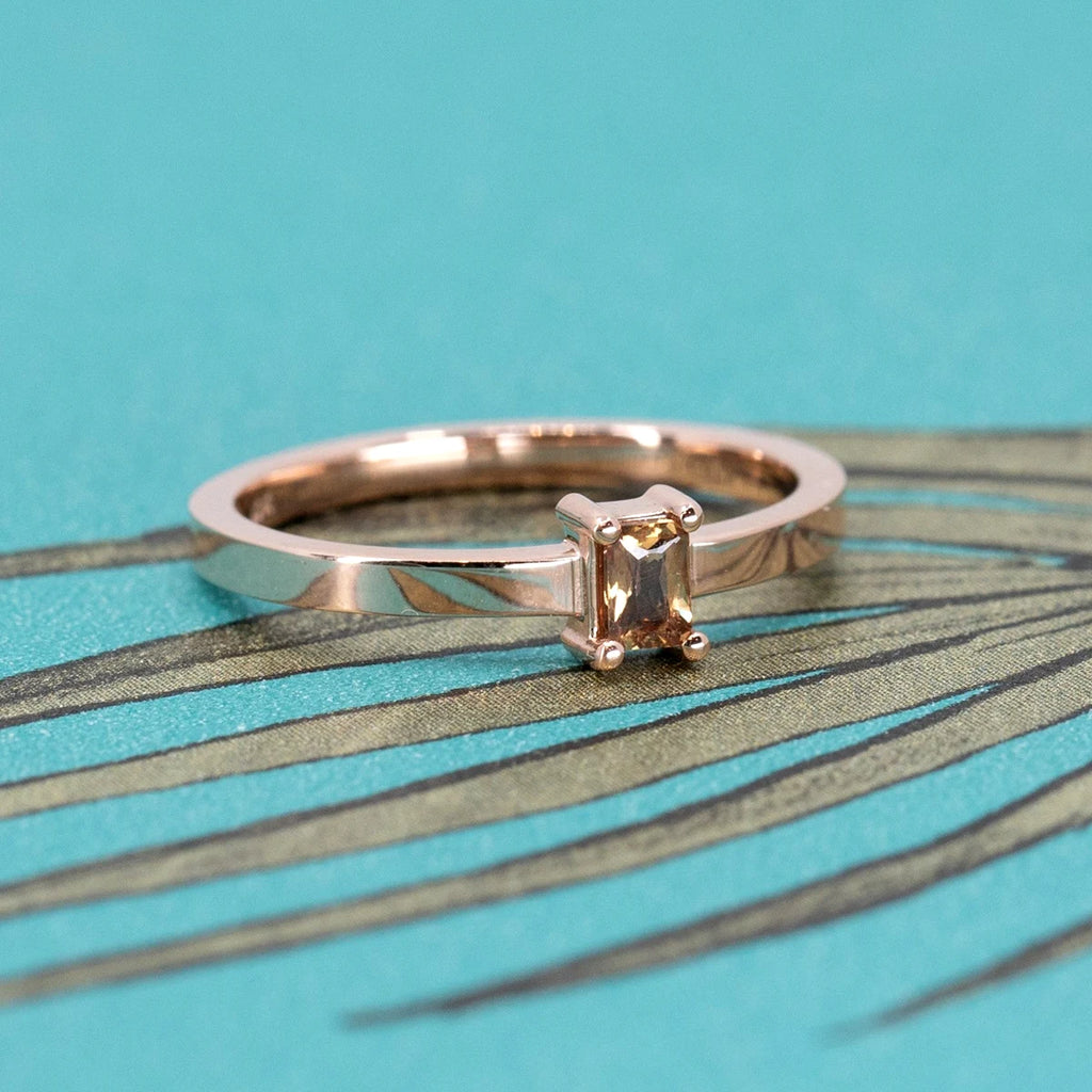 Minimalist rose gold ring featuring a golden-brown sapphire baguette. Photographed on a vibrant teal and gold background. Designed by Ruby Mardi.