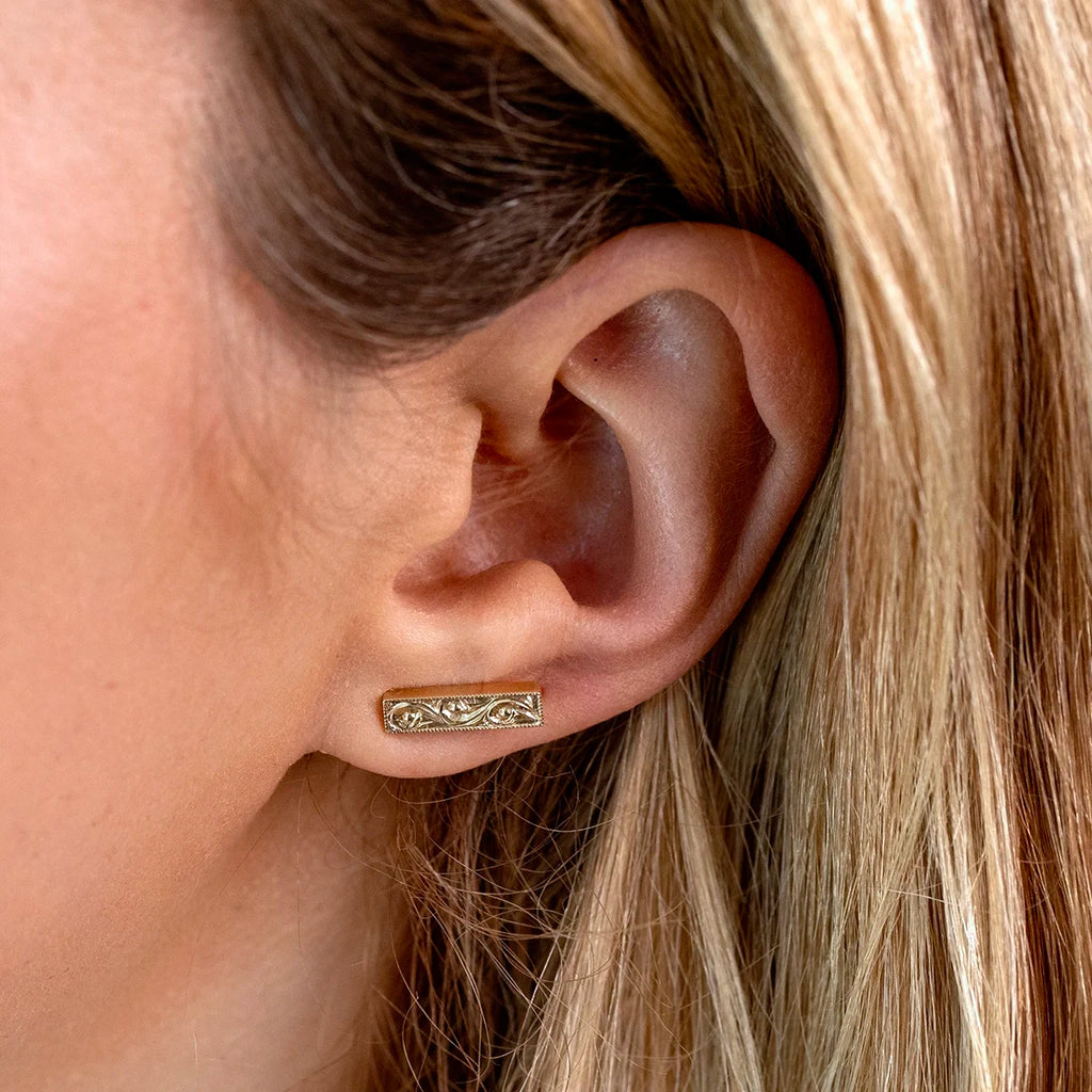 Close-up of a woman's ear wearing a rectangular 14k yellow gold stud earring with fine hand-engraved floral motifs and milgrain detailing along the edges. Jewelry by Ruby Mardi, handcrafted in Canada.