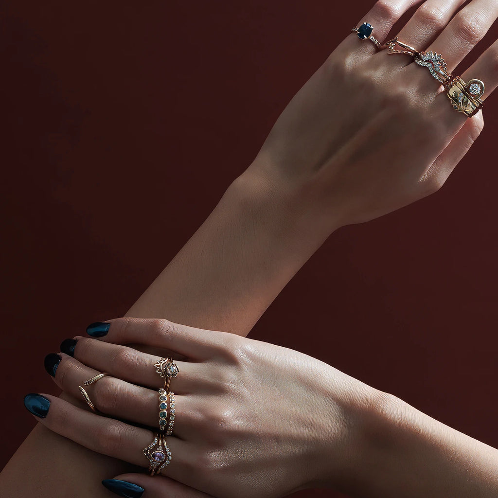 Two elegant hands photographed in studio on a wine color background. Many designer rings are stacked on the fingers. Alternative engagement rings, right hand rings and diamond wedding bands, all available at independent jewelry store Ruby Mardi.