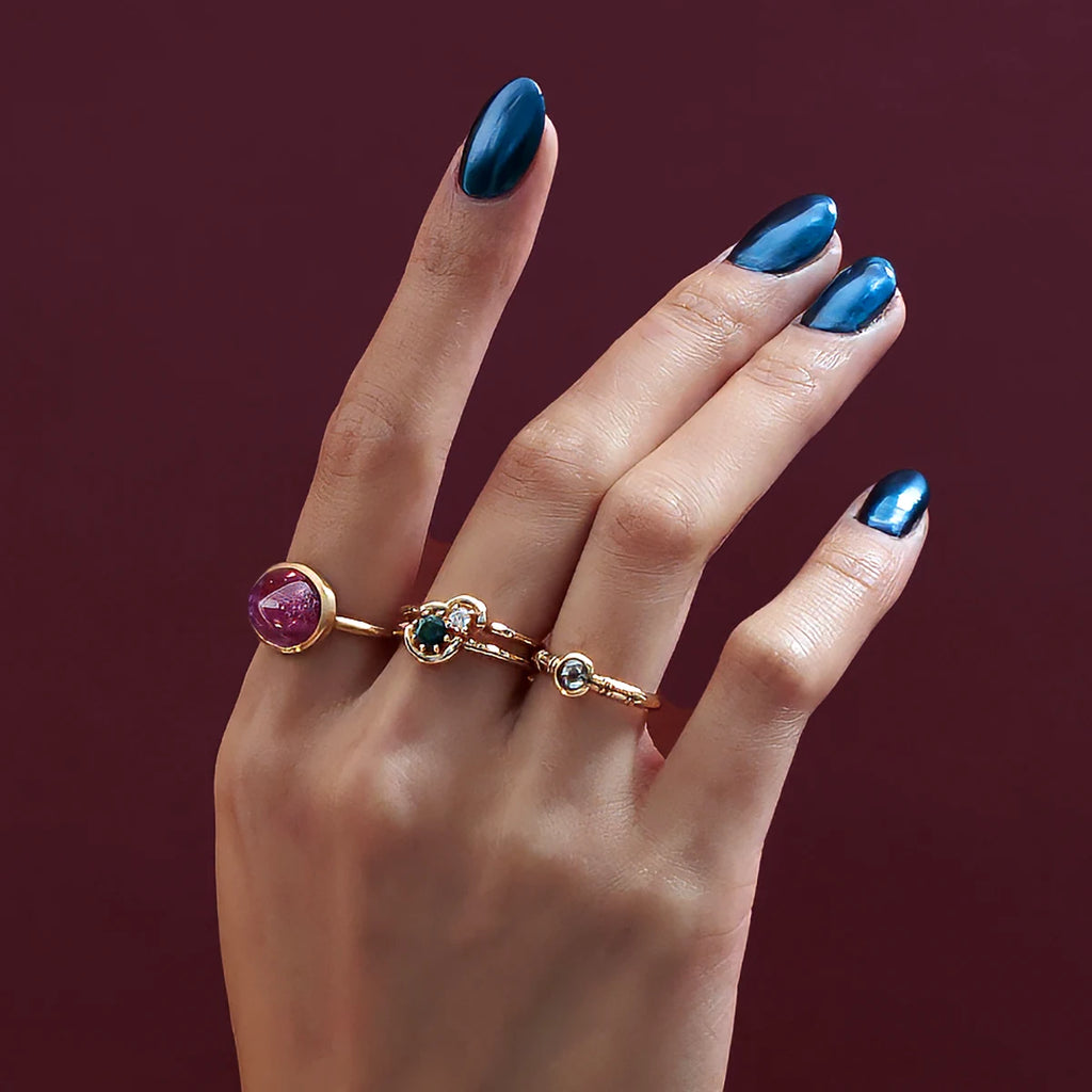 A hand with blue metallic nails is photographed in studio on a dark raspberry background. The hand wears 3 Canadian designer rings : one is a big ruby cabochon, one is with a salt and pepper diamond, and the one on the middle finger has a double yellow gold band and a teal sapphire as well as a diamond. These unique rings are available at the independent Montreal jewelry store Ruby Mardi.