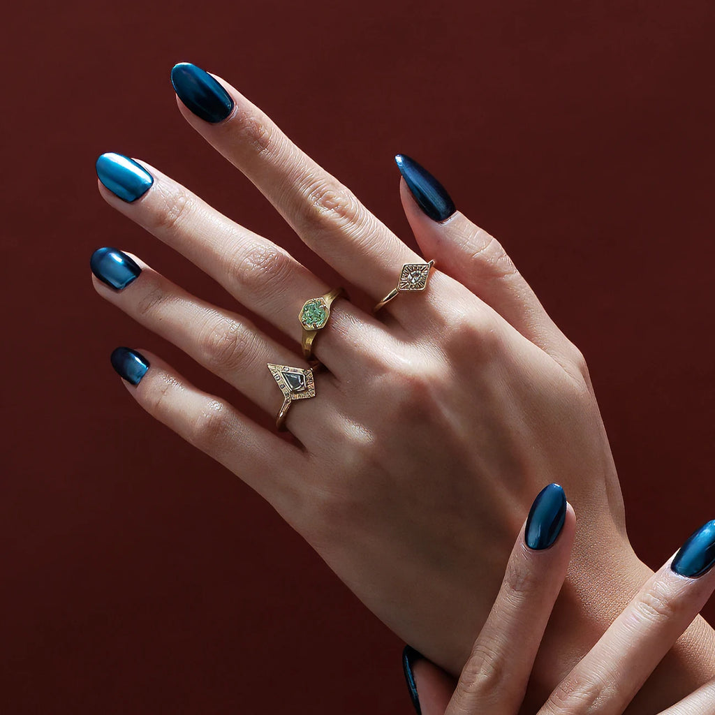 An artistic photography showing the hands of a lady that wears three designer rings. One has a kite shape and features a salt & pepper diamond. The other a raw green garnet set in yellow gold. The last a white sapphire and hand engraved details. The hand has electric blue nails. It's a picture from Canadian jewelry gallery Ruby Mardi, the best jewellery store in Canada.