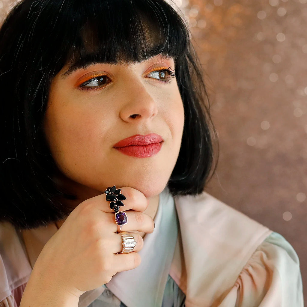 A woman with dark bobbed hair and warm-toned makeup gazes thoughtfully. Her hand rests under her chin, adorned with multiple statement rings, including a bold black gemstone ring and a faceted purple amethyst ring. The background is softly blurred with a warm, glittery effect, enhancing the elegance of the jewelry.
