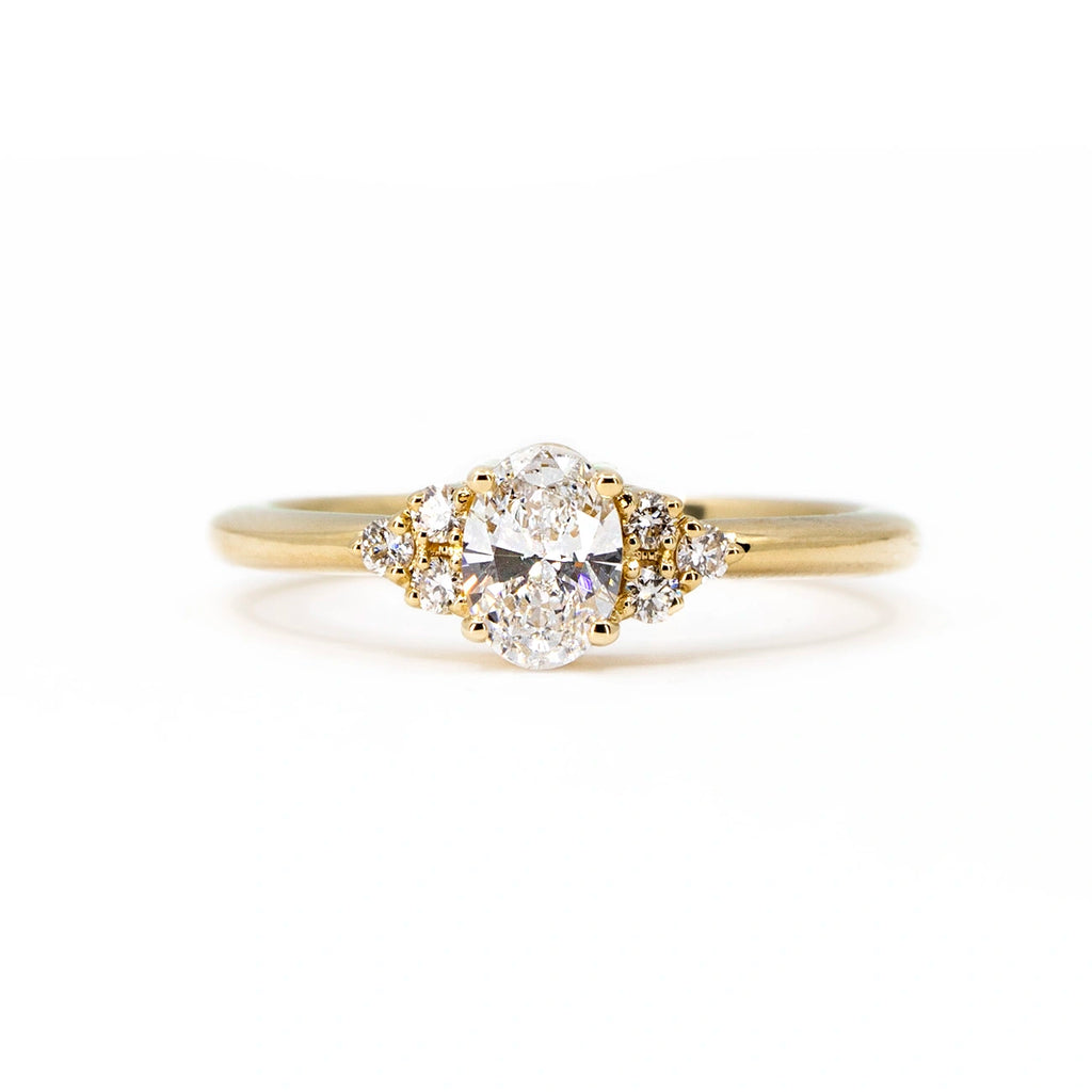 Classic engagement ring featuring a lab grown diamond in an oval shape and several small side diamonds. Custom made bridal ring designed by Ruby Mardi, a fine jewelry store that sells designer jewelry. The ring is photographed on a white background.