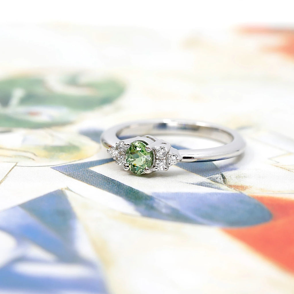 A classic white gold engagement ring featuring an oval green gemstone and diamond accents is photographed on funky artistic background. This bridal piece of jewelry with a garnet (January birthstone) was designed and handmade in Montreal by Ruby Mardi, a fine jewellery boutique that sells one-of-a-kind designer jewels.