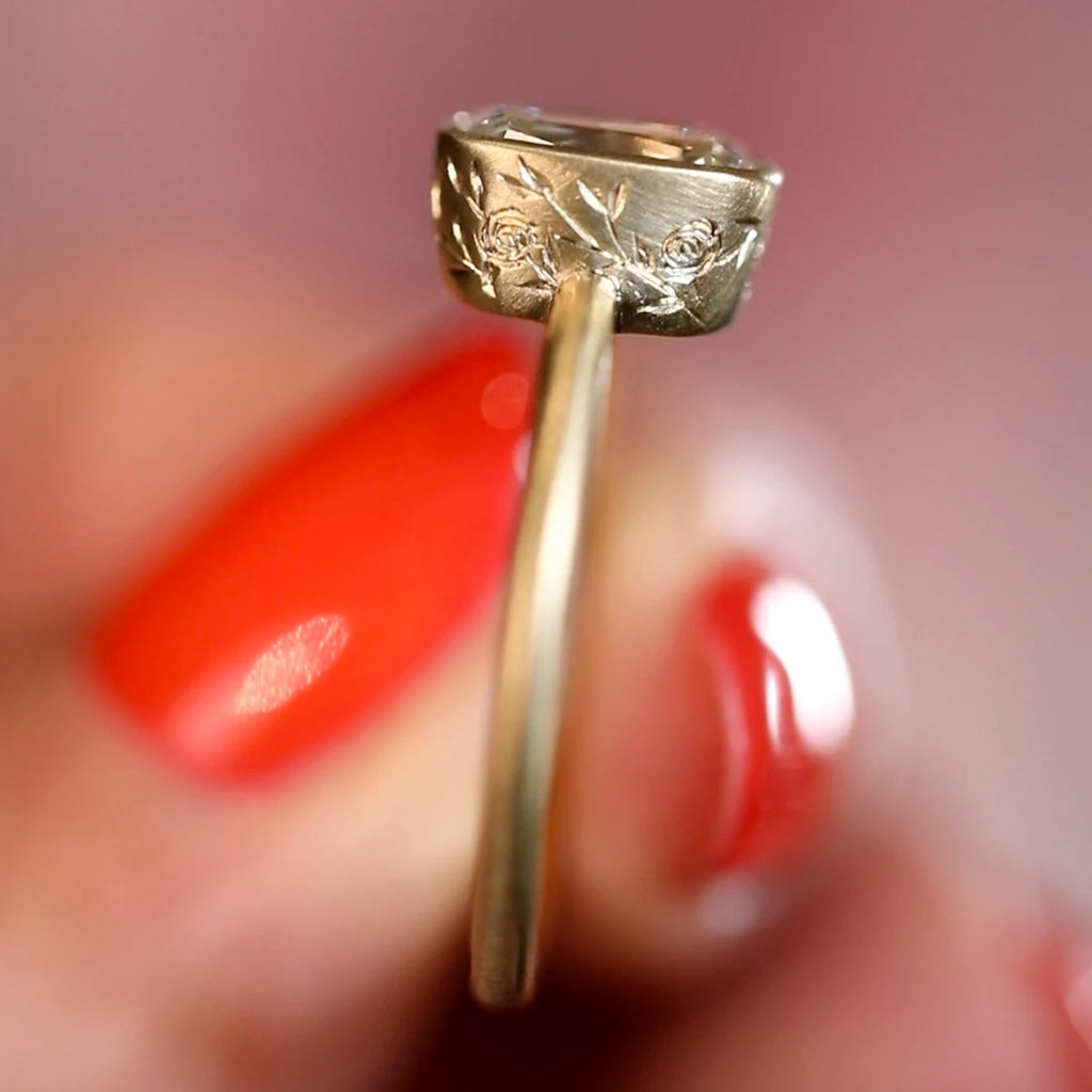 Close-up view of the engraved floral bezel of a handmade diamond solitaire ring in Fairmined matte yellow gold, crafted by Fluid Jewellery in Ottawa, Canada. Shown held between red-manicured fingers. Available at Ruby Mardi.