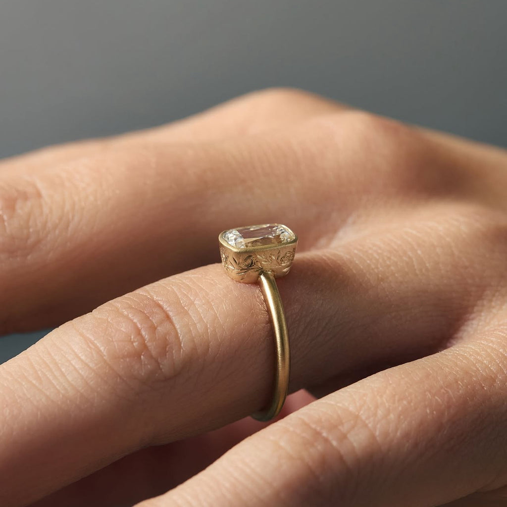 Close-up of a hand wearing a rectangular diamond solitaire ring with matte yellow gold band and delicate hand engravings around the bezel. Contemporary fine jewelry available at Ruby Mardi, handcrafted in Montreal.