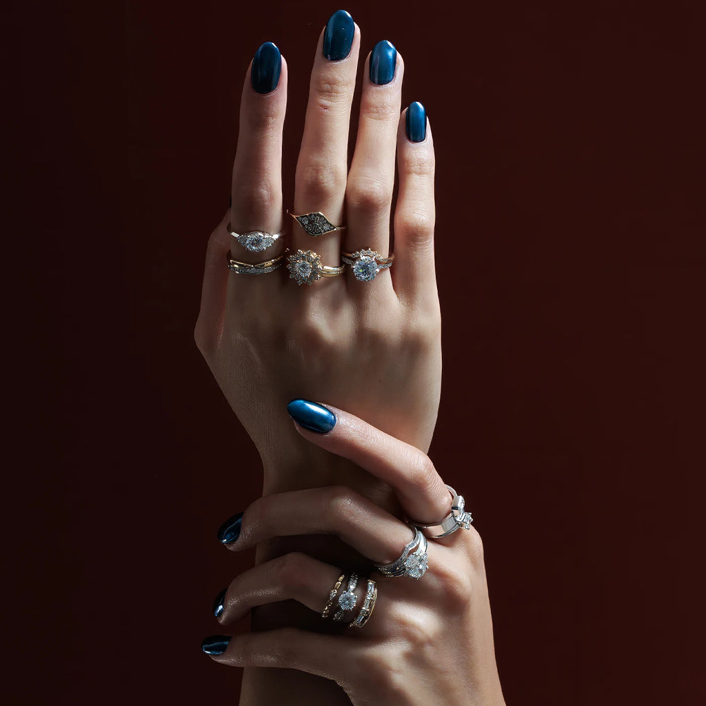 Two elegant hands, their nails painted metallic blue, wear designer diamond rings. The rings are all unique, different from the classic designs. They are handcrafted in Canada and available at the country's coolest jewelry store, Ruby Mardi.