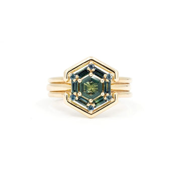 A ring with a unique design is photographed against a white background: the ring is in yellow gold and offers a 2-in-1 design: a wedding band fits inside the main ring, which can also be used as an engagement ring. It features 13 natural sapphires in shades of blue and green (teal), the whole forming a hexagonal shape. Created by Vancouver-based Erica Leal. This fine jewel is available at Canadian fine jewelry store Ruby Mardi.