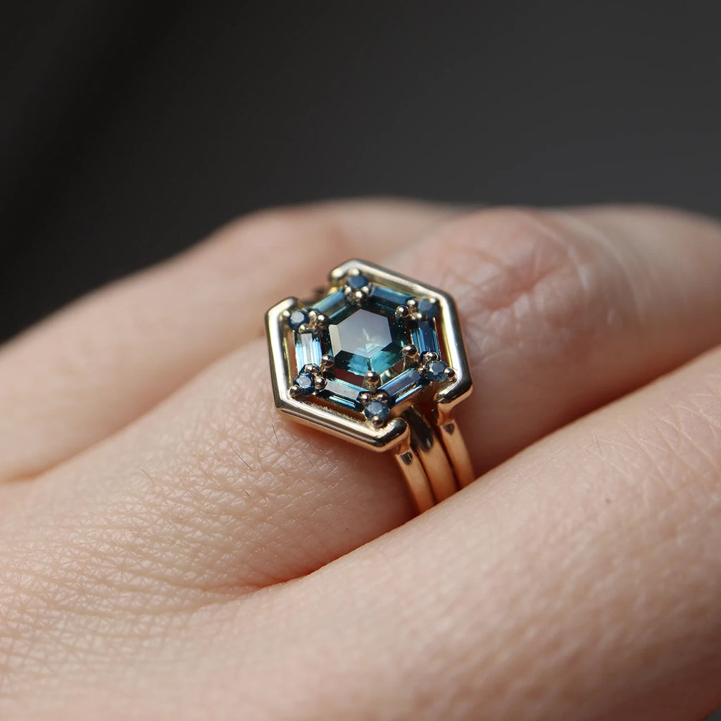 A close up on a hand of a yellow gold ring with a double band that features 13 teal sapphires of different shapes (round, baguette and hexagonal). This unique piece of fine jewelry is available at independent Canadian jewelry store Ruby Mardi.