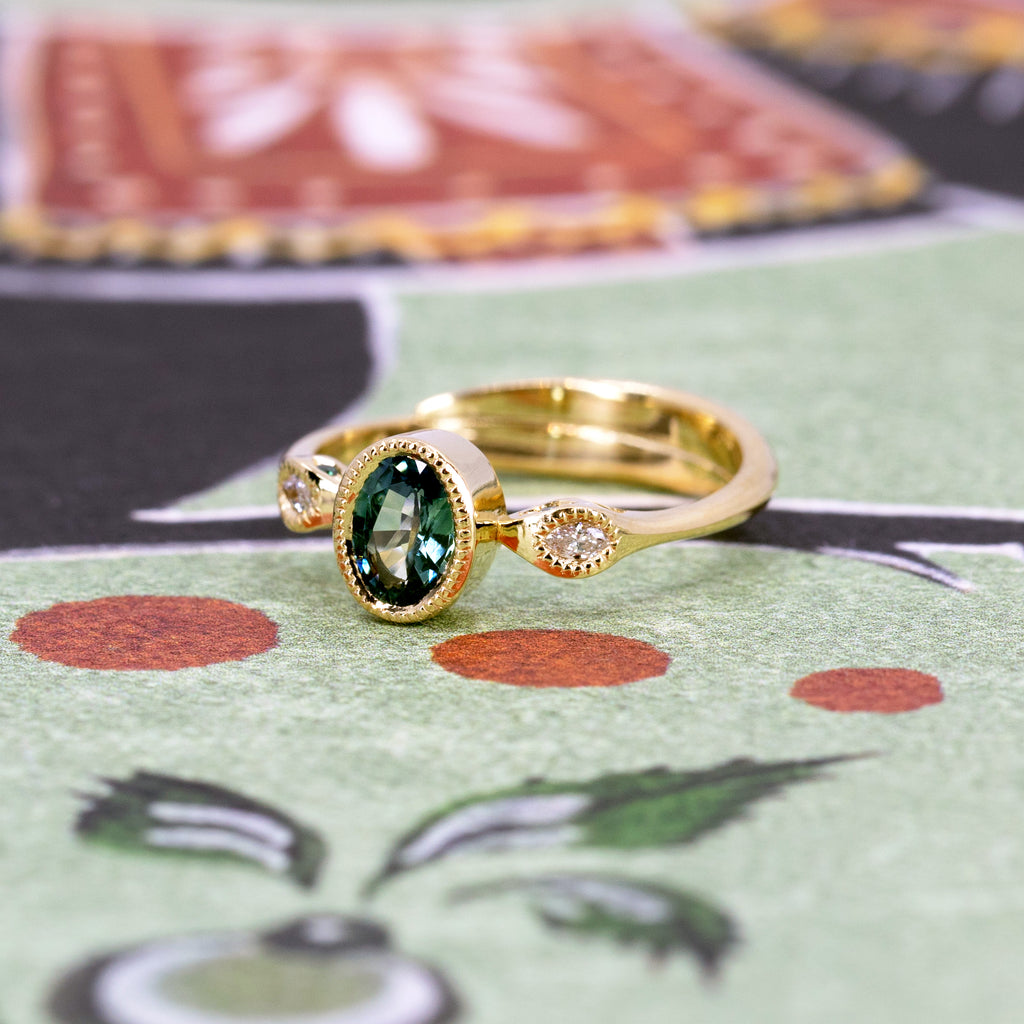 An absolutely unique yellow gold ring photographed against an abstract background: the band is formed by two superimposed snakes that join a central oval green sapphire on either side, set in a closed setting with miligrain detail. The gold snakes have white diamond eyes on the front and green diamond eyes on the sides! This exceptional engagement ring is available only at Canada's coolest jewelry store, Ruby Mardi. It was made by Emily Gill.