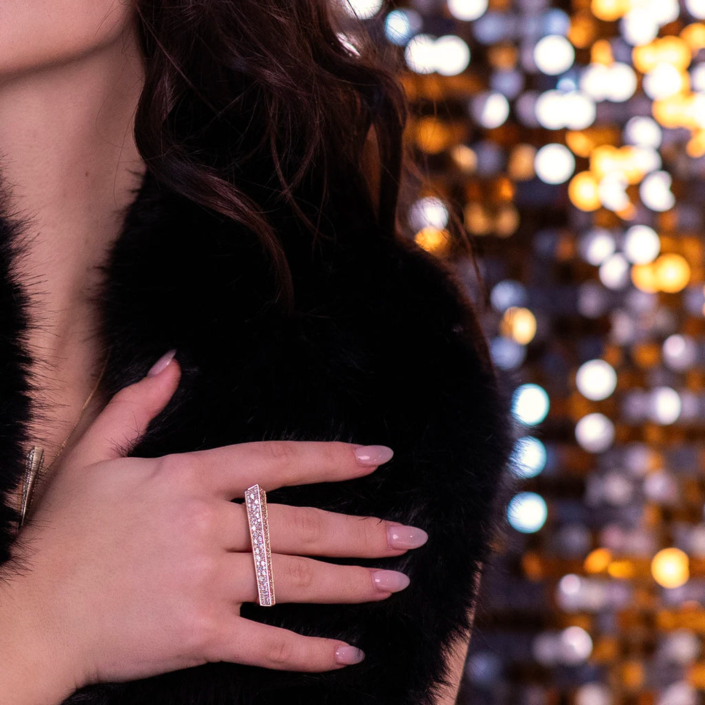 A glamorous close-up of the Punch ring, a bold double-finger design in gold, encrusted with radiant diamonds. Paired with a nude manicure and black fur, it exudes luxury and sophistication against a backdrop of warm golden lights.