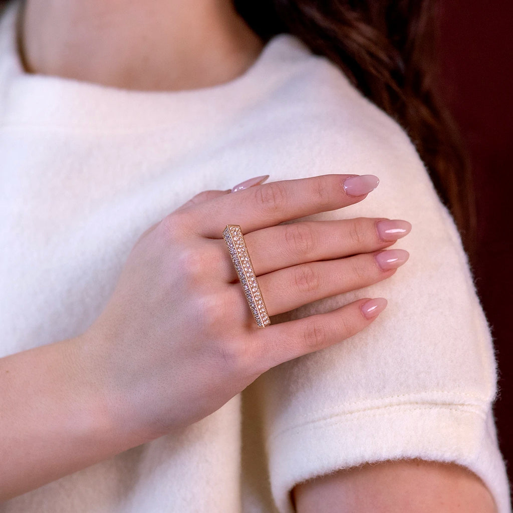 A close-up of a hand wearing the Punch double-finger ring, a bold and elegant design in gold encrusted with sparkling diamonds. The soft nude manicure and cream-colored outfit create a refined and minimalist backdrop, highlighting the striking statement piece.