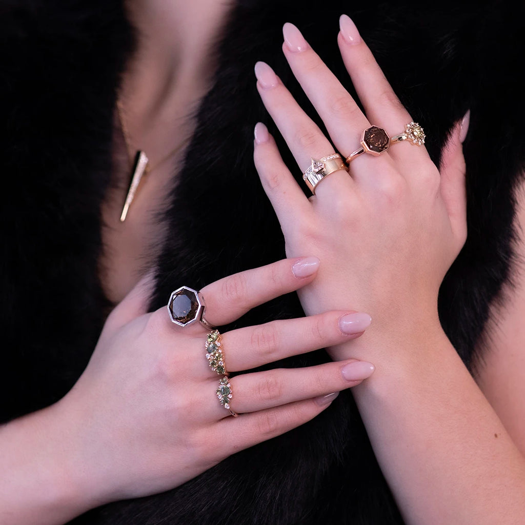 A close-up image of a person's hands adorned with multiple gold rings, including sapphire cluster rings, a modern gold wedding set, and a large hexagonal smoky quartz set in white gold. The rings showcase a mix of contemporary and classic designs. The person is dressed in a black fur-like garment and wears a gold pendant necklace, contributing to the luxurious and refined atmosphere of the image.
