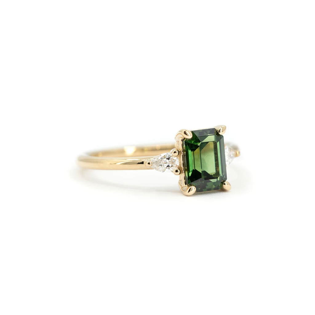 Side view of a yellow gold engagement ring with an emerald-cut green sapphire and pear-shaped white diamond accents. Handcrafted by Maksym Jewelry in Montreal, available at Ruby Mardi.