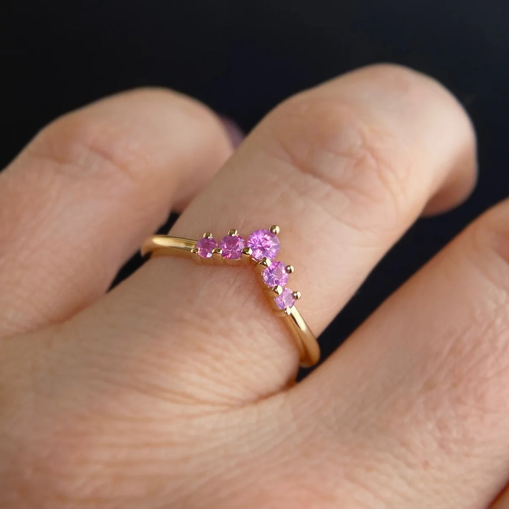 Shimmerette crown-shaped pink sapphire wedding band worn on finger. Featuring five luminous pink sapphires in a graceful V contour, set in 14k yellow gold. This delicate stacking ring is handcrafted by Canadian jewelry designer Emily Gill for Ruby Mardi in Montreal. Ideal for pairing with engagement rings or worn solo for a colorful statement.