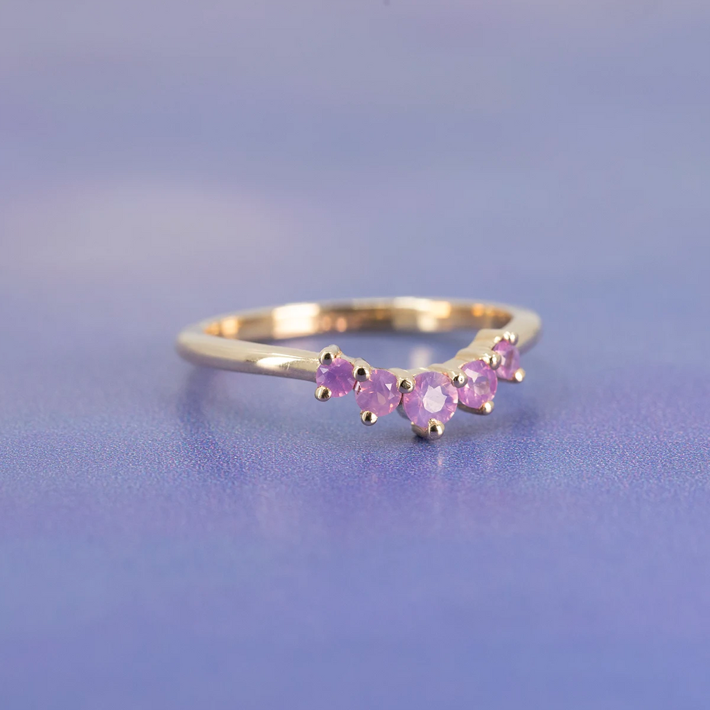 Bagatelle ring by Emily Gill featuring five heat-treated pink sapphires in a contour band design, set in 14k yellow gold. Photographed on a soft violet background. Handmade in Canada and available at Ruby Mardi, Montreal.